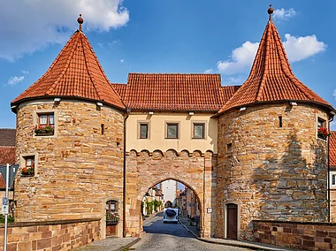 Ein historisches Stadttor mit roten Dächer und zwei runden Türmen an den Ecken gibt den Blick frei hinein in ein mittelalterliches Städtchen