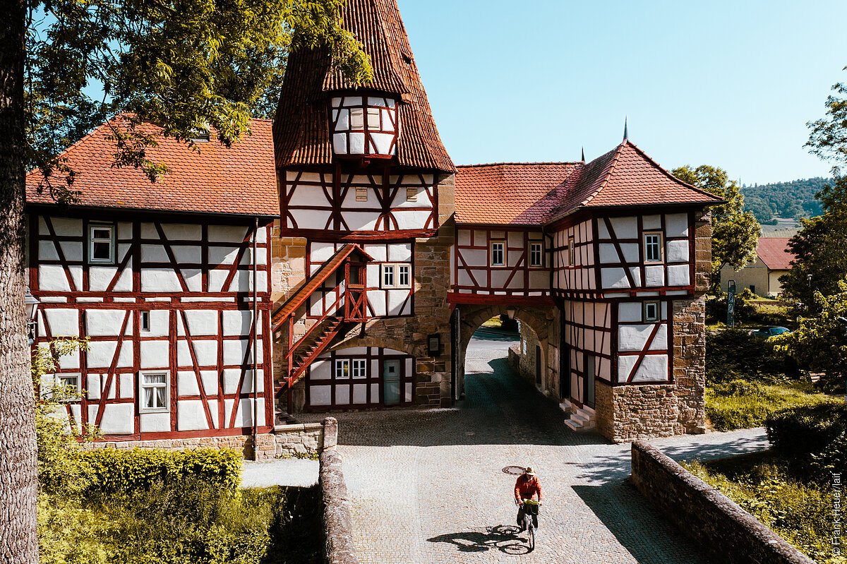 Fachwerkgebäude mit Torbogen und rotem Dach, Radfahrer auf gepflastertem Weg. Bäume im Hintergrund.