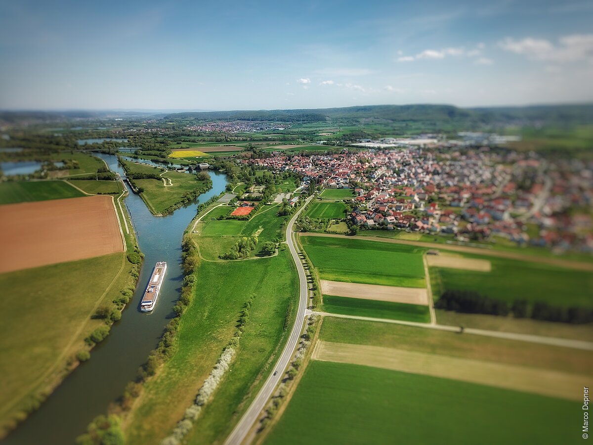 Ortsansicht (Knetzgau, Steigerwald) Luftaufnahme einer Flusslandschaft mit einem Frachtschiff, umgeben von Feldern und einer Siedlung im Hintergrund.